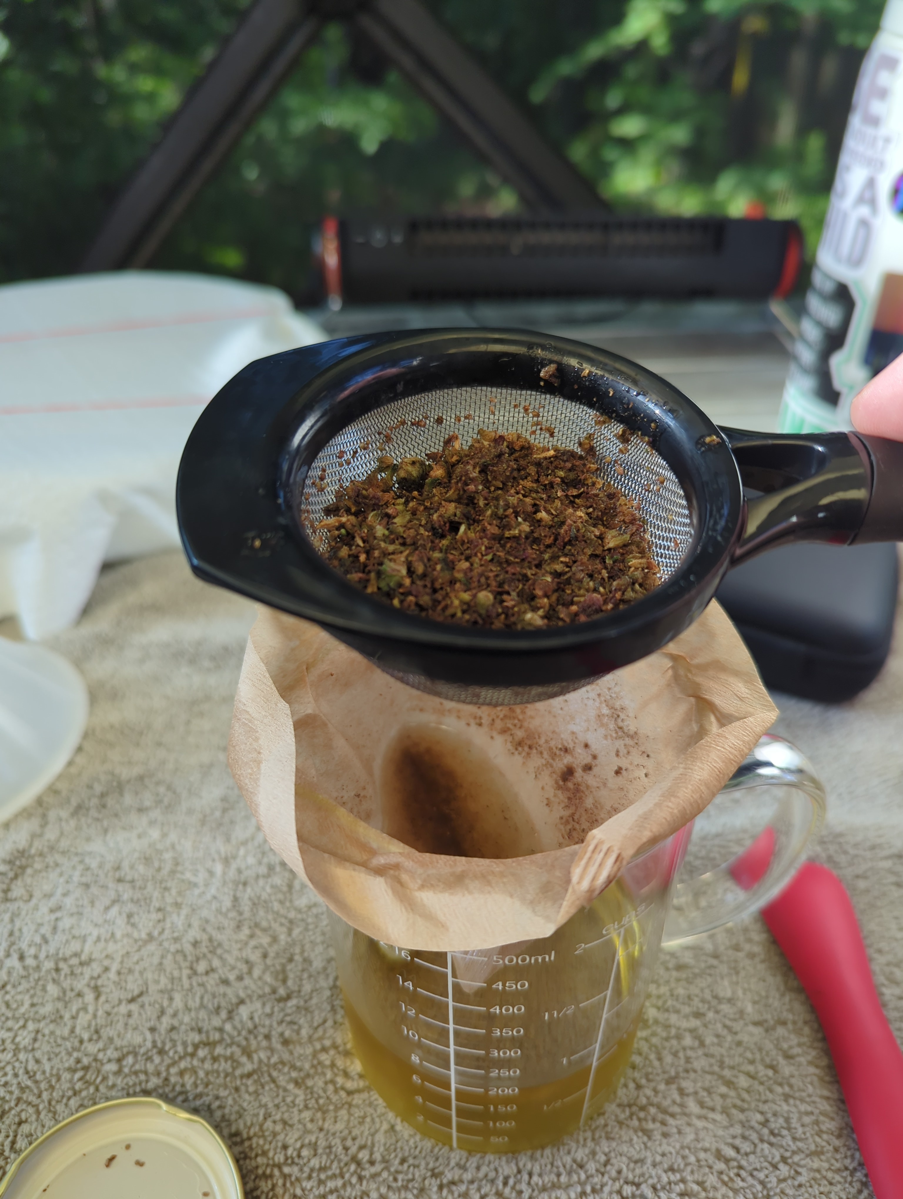 a glass beaker with an unbleached coffee filter in the top. a small tea strainer holds cannabis material over it. the beaker contains golden liquid